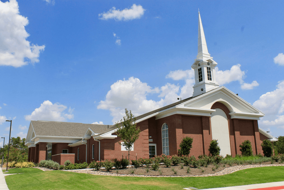 A red brick meetinghouse with a tall white steeple, behind a lawn and young trees under a bright blue sky.